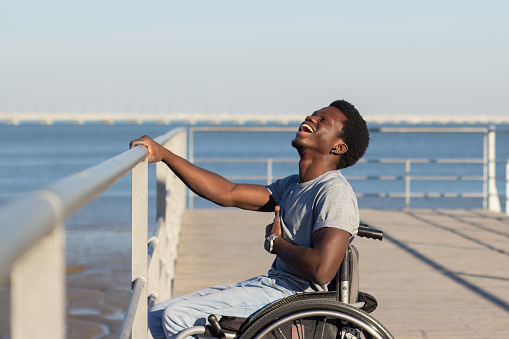 Joyful Black man sitting in wheelchair at seafront on sunny day. Side view of happy guy with disability holding railing with his hand, and laughing out loud. Disability, attitude concept.