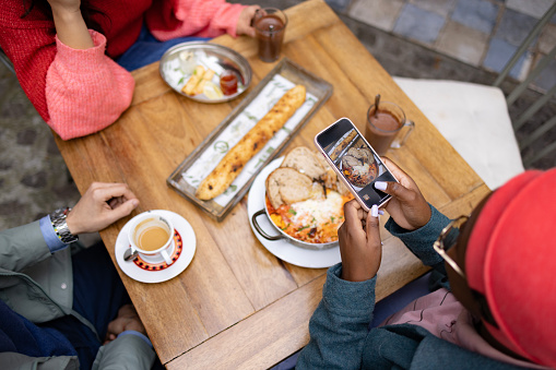 High angle view of a small group of friends sitting in the restaurant and having a lunch together, one of them using a smart phone to take a photograph of the food they ordered