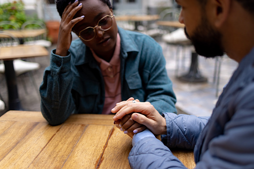 Sad young adult woman meeting with her male friend in a local coffee shop, telling him about the problems she is having while he is listening to her and holding her hand