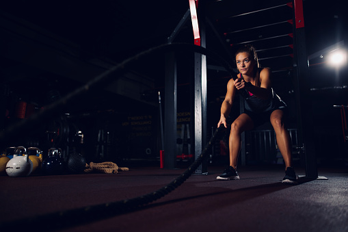 Young fit sportswoman working out with battle ropes at gym