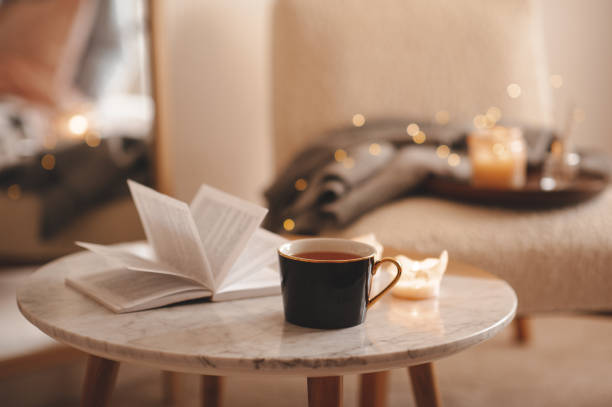 Cozy Christmas home atmosphere Cup of tea with paper open book and burning scented candles on marble table over cozy chair and glowing lights in bedroom closeup. Winter holiday season. hygge stock pictures, royalty-free photos & images