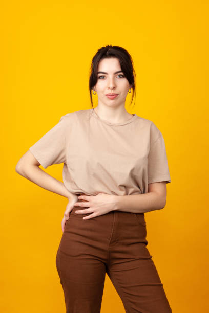 Studio portrait of a cheerful 20 year old white woman with black hair in a beige t-shirt and brown pants against a yellow background stock photo