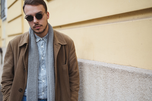 Young street fashion model with sunglasses,  brown coat and scarf posing against yellow textured wall, looking at camera