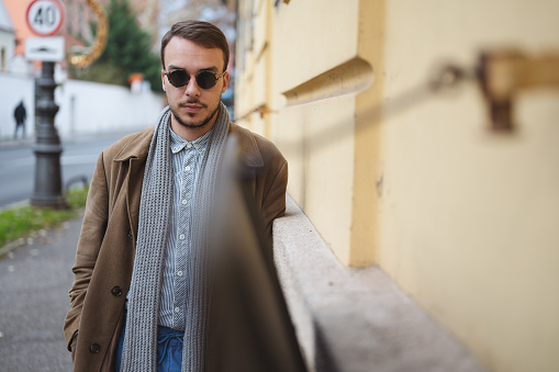 Young street fashion model with sunglasses and brown coat leaning on building wall, looking at camera