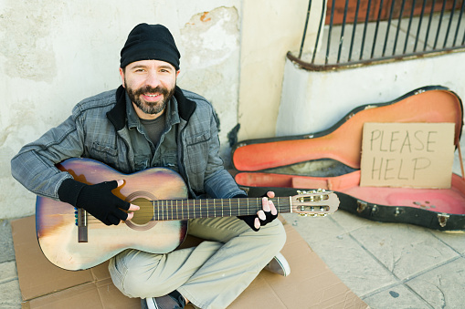 Happy poor homeless man smiling while playing the guitar while asking for help and money