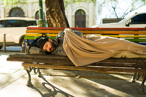 Dirty cold homeless man in poverty covered with a blanket sleeping alone on the park bench