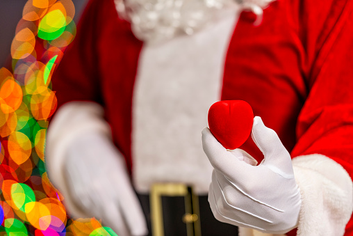 Santa Claus holding a red velvet heart shaped jewelry box with bokeh of multi-colored Christmas tree lights in the background