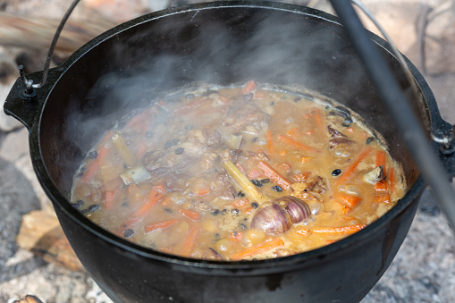 Fried lamb meat languishes in broth with vegetables in a cauldron on a campfire, cooking lunch for a large company at a campsite