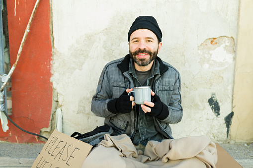 Happy homeless man living in the street smiling while drinking a cup of coffee while showing a please help cardboard sign