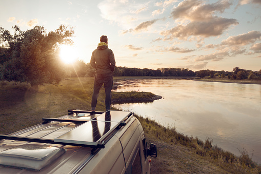 A man is enjoying the view from the rooftop of his camper van which is parked next to a river.
