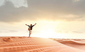 Hiker man walking in the desert sand dunes at sunset - Happy traveler with arms up enjoying freedom outside - Wanderlust, wellbeing, happiness and travel concept