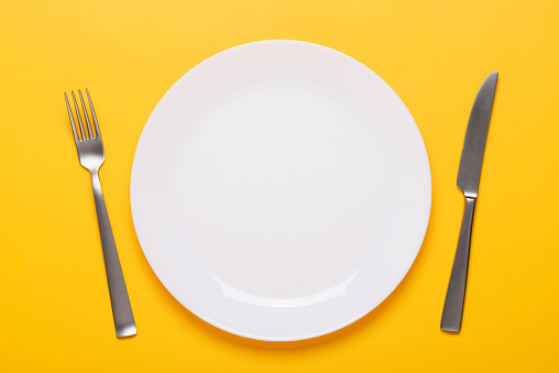 Clean empty white plate with knife and fork on yellow background place setting top view
