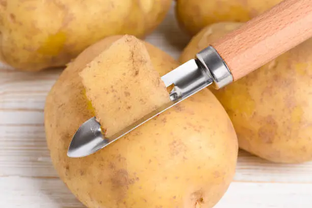 Raw potato peeled with vegetable peeler on white wooden table