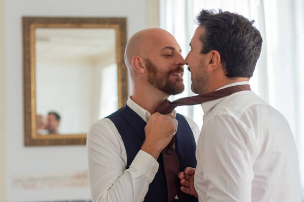 Gay holding partners tie and kissing him in hotel room Gay holding partners tie and kissing him in hotel room. Excited homosexual couple getting ready for wedding ceremony, flirting, enjoying sweet moments. LGBT, love, marriage concept gay wedding kiss stock pictures, royalty-free photos & images