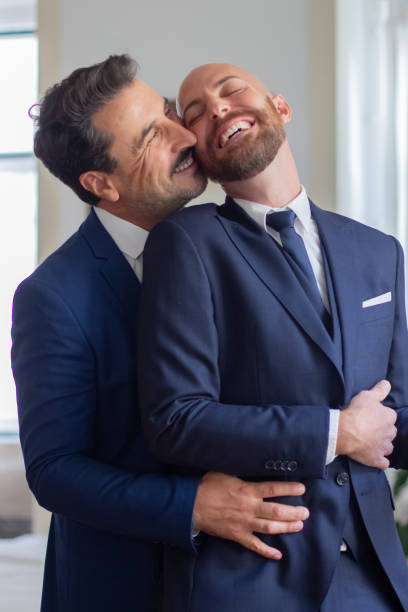 Happy gay in official suits hugging and kissing in hotel room Happy gay in official suits hugging and kissing in hotel room. Vertical shot of handsome homosexual couple laughing, getting dressed for wedding ceremony, having fun together. LGBT, love concept gay wedding kiss stock pictures, royalty-free photos & images
