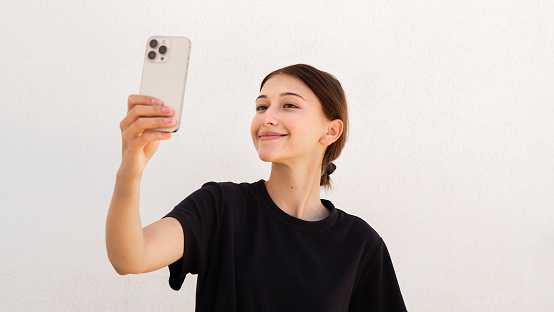 Portrait of happy young woman looking at cellphone as in mirror over white background. Caucasian woman wearing black T-shirt taking selfie with mobile phone. Mobile technology concept