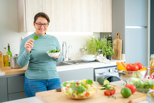 Healthy eating: smiling hispanic woman holding a spinach leaf while preparing fresh vegetables salad in domestic kitchen. High resolution 42Mp indoors digital capture taken with SONY A7rII and Zeiss Batis 40mm F2.0 CF lens