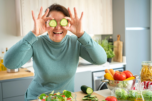 Healthy eating: smiling hispanic woman holding cucumber slices in front of her face while preparing fresh vegetables salad in domestic kitchen. High resolution 42Mp indoors digital capture taken with SONY A7rII and Zeiss Batis 40mm F2.0 CF lens