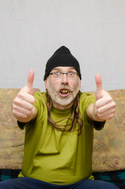 Ugly man with bad teeth (false dentures) at home during covid-19 pandemic lockdown and making two thumbs up.He wears a tuque over his long hair and a beard