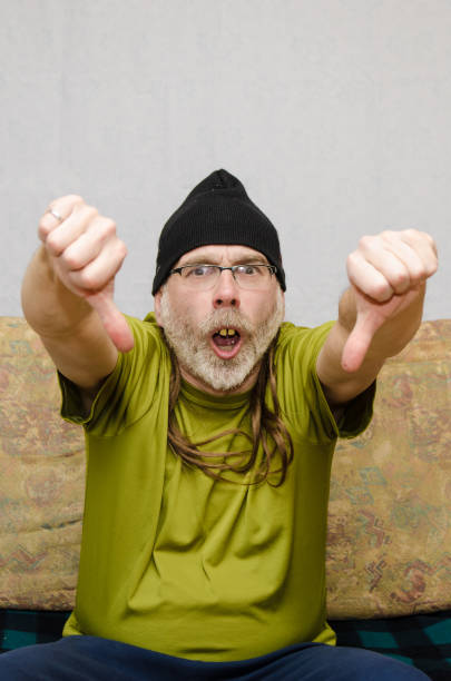 Ugly man with bad teeth (false dentures) at home during covid-19 pandemic lockdown and making two thumbs down.He wears a tuque over his long hair and a beard