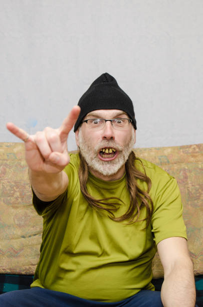 Ugly man with bad teeth (false dentures) at home during covid-19 pandemic lockdown and making two horn sign.He wears a tuque over his long hair and a beard