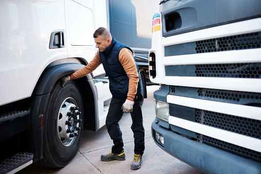 Mid adult trucker standing outside his semi-truck is checking the wheel under the cab on the tractor before hitting the road. Full length, copy space