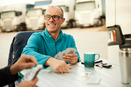 Two truckers, one partially viewed and the other with a big smile, outdoors playing cards at a rest area. Close-up