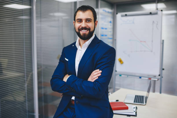 retrato de media longitud de exitoso jefe masculino vestido con elegante traje con los brazos cruzados sonriendo a la cámara durante el tiempo de trabajo, hombre experimentado en ropa formal disfrutando de estilo de vida de negocios en el interior - economist fotografías e imágenes de stock