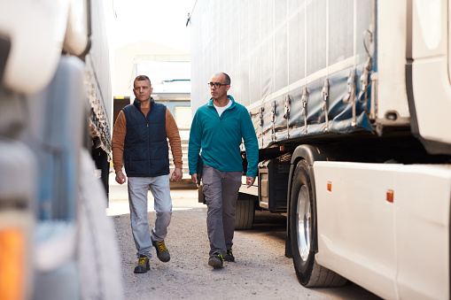 Two male truckers walking and talking between two semi-trucks. Full length