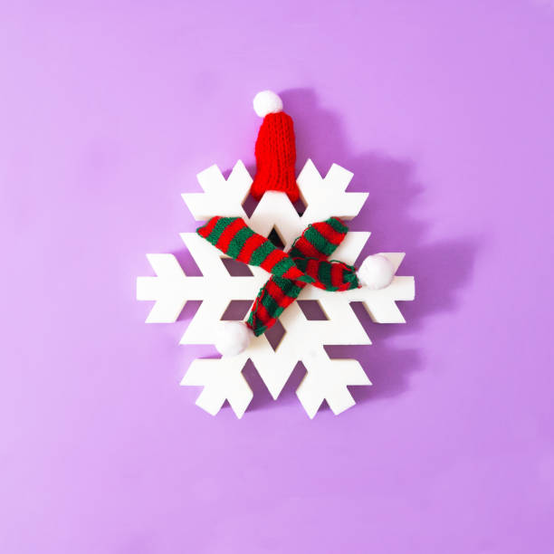 A big white snowflake with red and green striped winter scarf and a red hat, on lilac background. Minimal surreal concept for season greetings card or banner. Design for season greetings card or banner un gran copo de nieve blanco con bufanda de invierno a rayas rojas y verdes y un sombrero rojo, sobre fondo lila. concepto surrealista mínimo para tarjeta de felicitación de temporada o banner. diseño para tarjeta de felicitación o banner de temporada