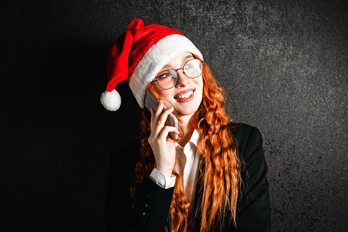 Portrait of a red-haired business woman in a suit with a smartphone in a red Christmas hat.