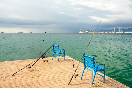 2-3 fishing rods and blue chairs on the pier and bay of Cannes, as well as a lighthouse and a cruise ship in the distance and yachts anchored in the water of the Mediterranean Sea on the French Riviera