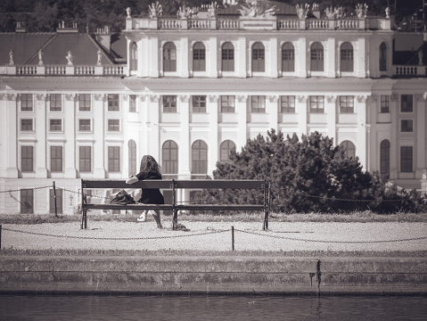 Vienna, Austria - June 2022: Rear view with a girl sitting on a bench in the gardens of Schonbrunn Palace Vienna, Austria. Black and white street photography.