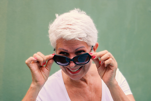 Stylish old lady with white hair, white t-shirt and sunglasses against green background