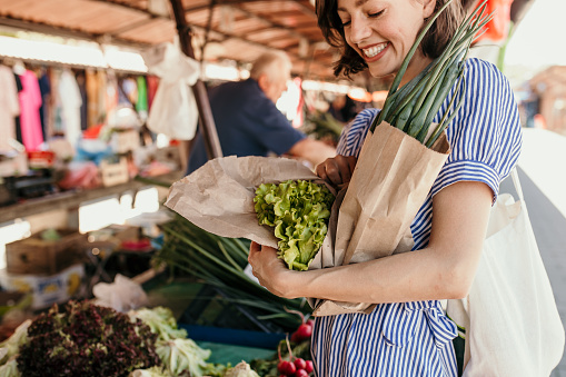 A smiling young lady is standing by a stand with fresh vegetables. She is holding various vegetables and she decided to buy everything and put it in a reusable bag.