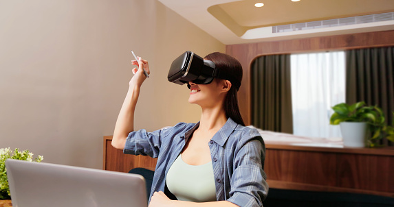 asian happy woman wearing virtual reality glasses with vr technology to learn online education by laptop computer in hotel room or at home - she use stylus pen tap and write on air remoting