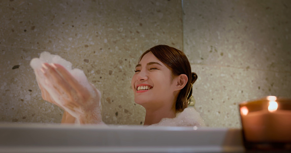 happy asian woman enjoy take a bubble foam bath inside bathtub with scented candle in bathroom - she play and blow foaming on her hand