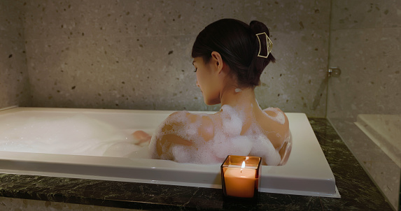 Smiling cheerful woman in towel brushing leg next to big ceramic bathtub, making anti-cellulite or lymphatic drainage massage on hips stepping on wooden stool in luxury bathroom. Body care, spa