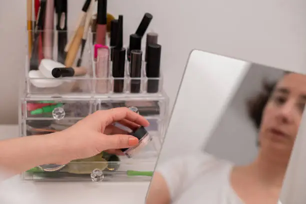 A woman is taking out a jar from a transparent make-up organiser’s drawer, woman’s face is seen from the reflection on the mirror.