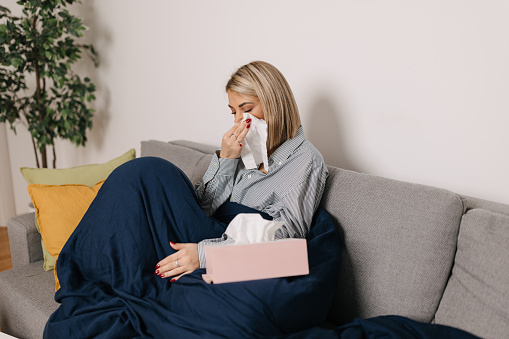 A young Caucasian woman has a cold and is sitting on her living room sofa, blowing her nose.