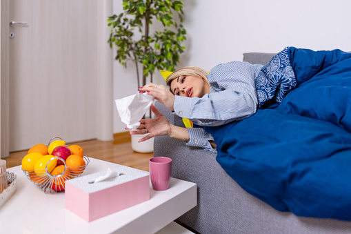 A young Caucasian woman is lying on her living room sofa sick and taking napkins from a box on the table.