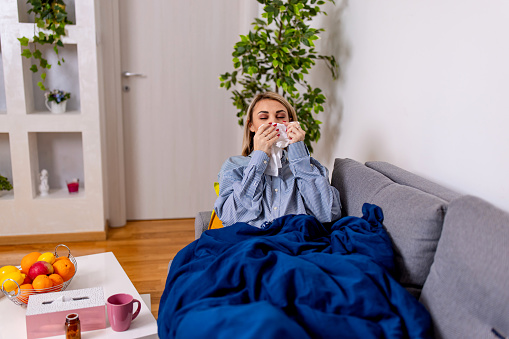 A young Caucasian woman is lying sick on her sofa under a blanket, and blowing her nose using a napkin.