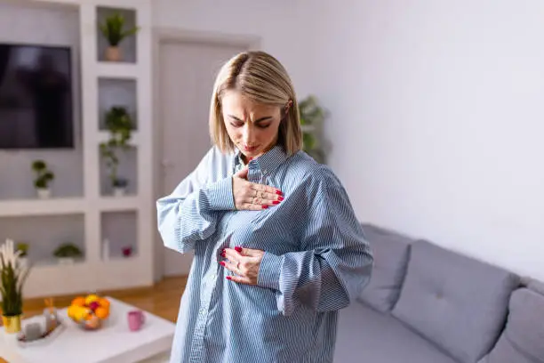 A young Caucasian woman in casual clothes is standing in her living room and checking her breasts for possible lumps.