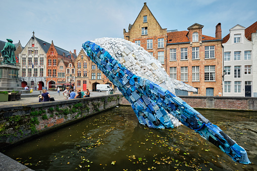 Bruges, Belgium - May 29, 2018: Skyscraper or The Bruges Whale - whale rising up from the Canal installation made up of 5 tons of plastic waste pulled out of the ocean in front of Jan van Eyck statue