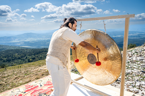 Musician playing bronze gong with drumsticks, mountain landscape in background. Music therapy, stress relief, mental health, harmony concept
