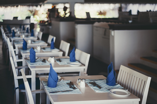 An indoor resort canteen with many made tables prepared for lunch served with blue rag napkins and cutlery; white wooden chairs, and tables; shallow depth of field, selective focus on the first table