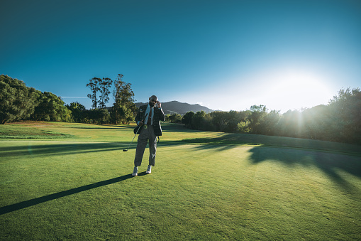 A wide-angle view of a dashing bald African American man in an elegant tailored outfit adjusting his sunglasses while staying on the lawn of a golf field with a club in his hand on a sunset