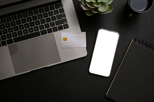 Stylish black tabletop workspace with smartphone white screen mockup, credit card, laptop computer, notepad and decor. top view, flat lay