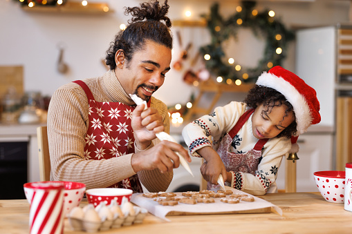 Happy african american family father and son in aprons making Christmas cookies together while cooking in kitchen at home, smiling   boy helping dad to decorate xmas gingerbreads during winter holidays