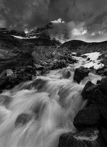 A black and white image shows a raging river pouring over rocks in the Italian Alps. The famous Matterhorn peak is seen in the background, shrouded in dramatic clouds.